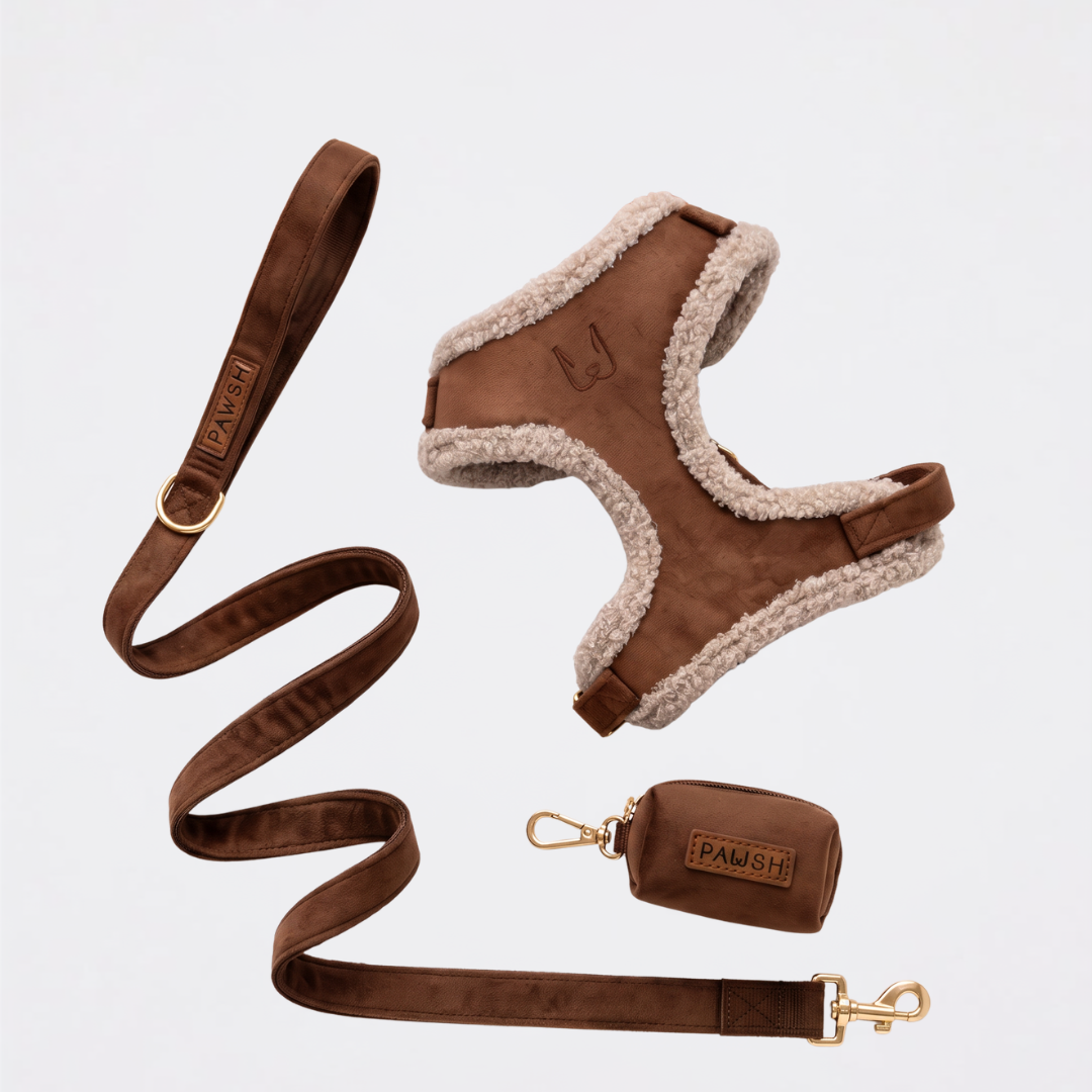 A dog harness set in brown suede with a cream sherpa lining, accompanied by a matching brown leash, a small brown leather pouch, and gold-tone hardware, all laid out on a light gray background